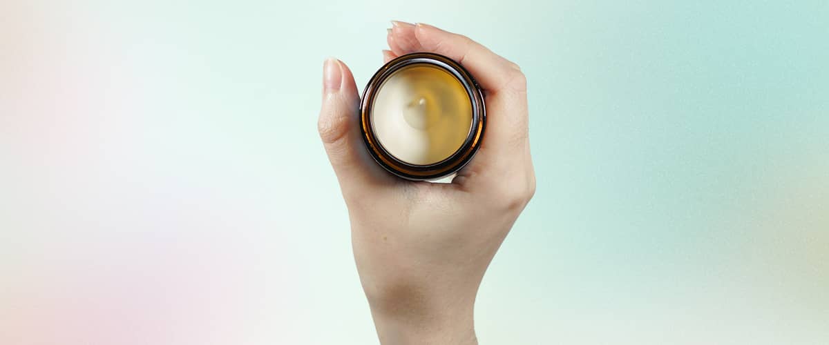 Mână feminină ținând un borcan maro de cremă deschis, vedere de sus în care se vede crema albă-bej din interior fără capac, fundal degradat pastel verde-roz