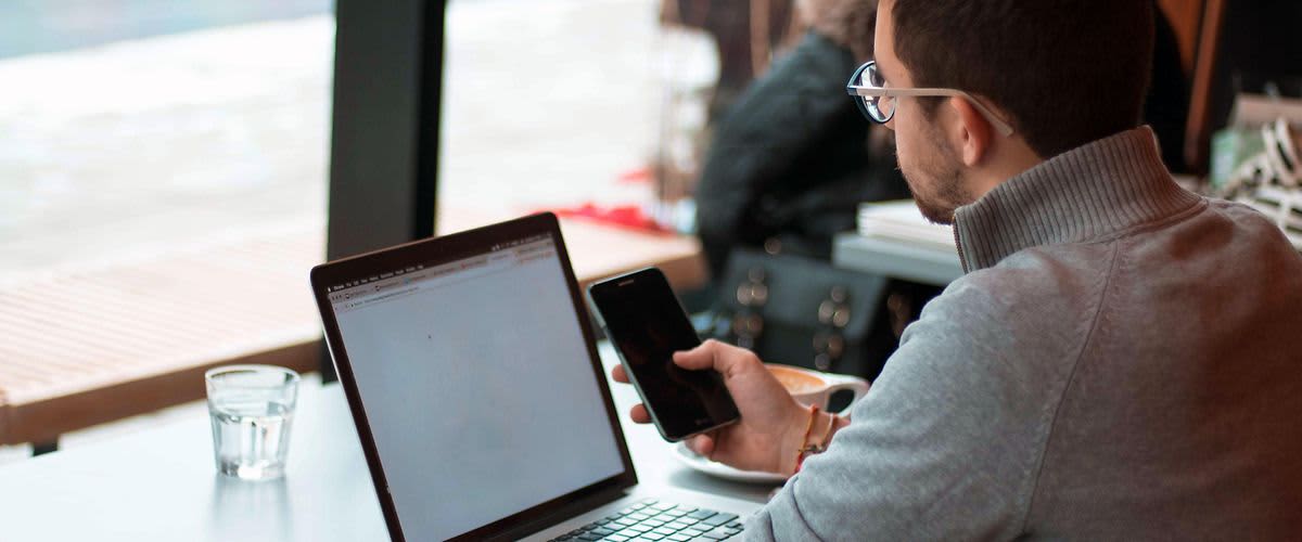 Un bărbat se află în fața laptopului său și formează un număr pe telefonul său mobil