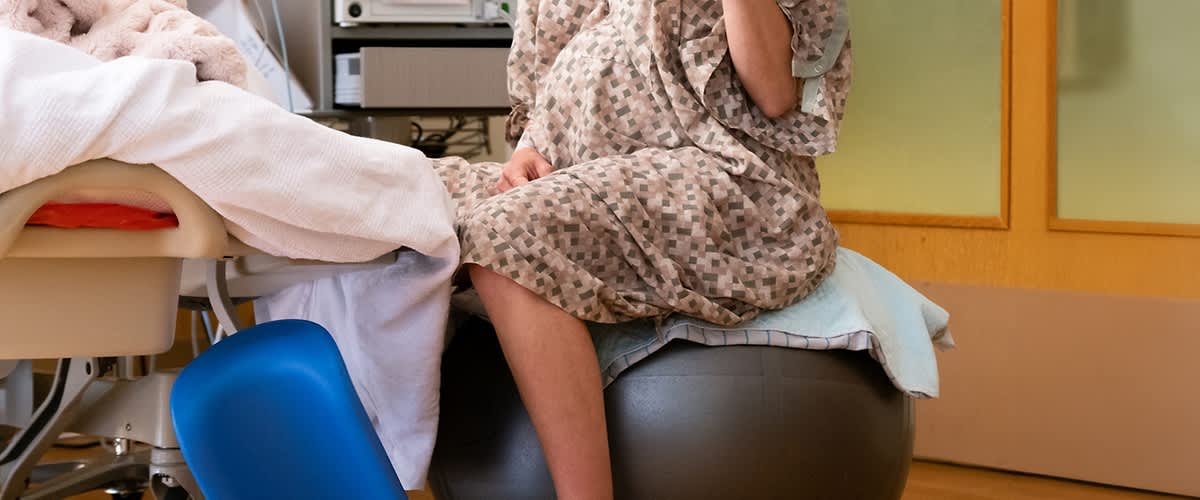 Femeie gravidă în halat stând pe minge de exerciții lângă pat de spital
