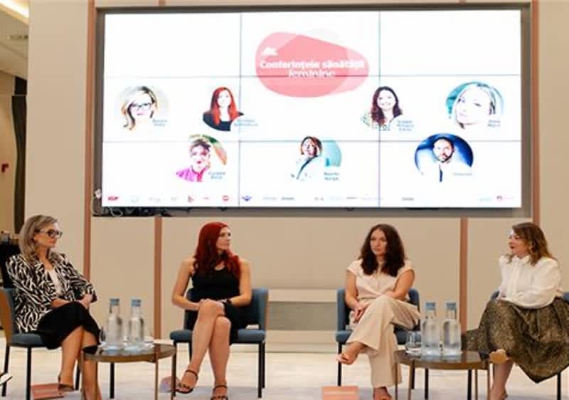 Patru femei stând pe scaune în fața unui ecran mare la o conferință