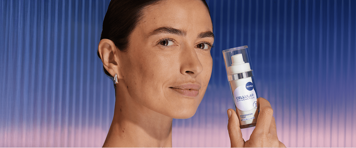 Mână ținând o cremă anti-îmbătrânire Nivea în fața unui fundal albastru și roz