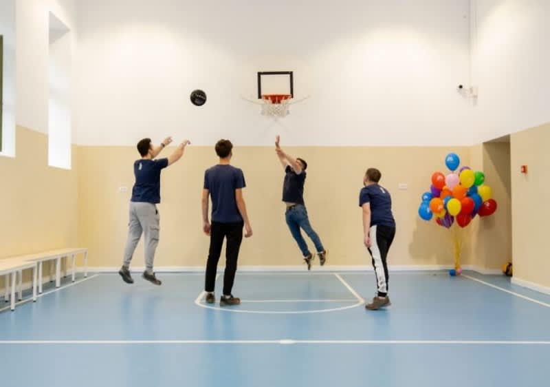Patru tineri joacă baschet într-o sală, cu baloane colorate în fundal