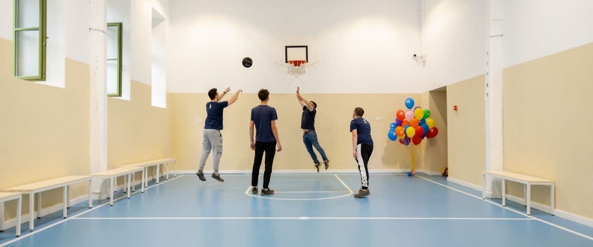 Patru tineri joacă baschet într-o sală, cu baloane colorate în fundal