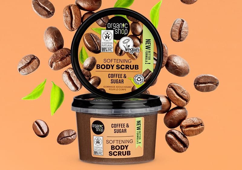 Recipient de scrub de corp cu cafea și zahăr, înconjurat de boabe de cafea și frunze verzi.