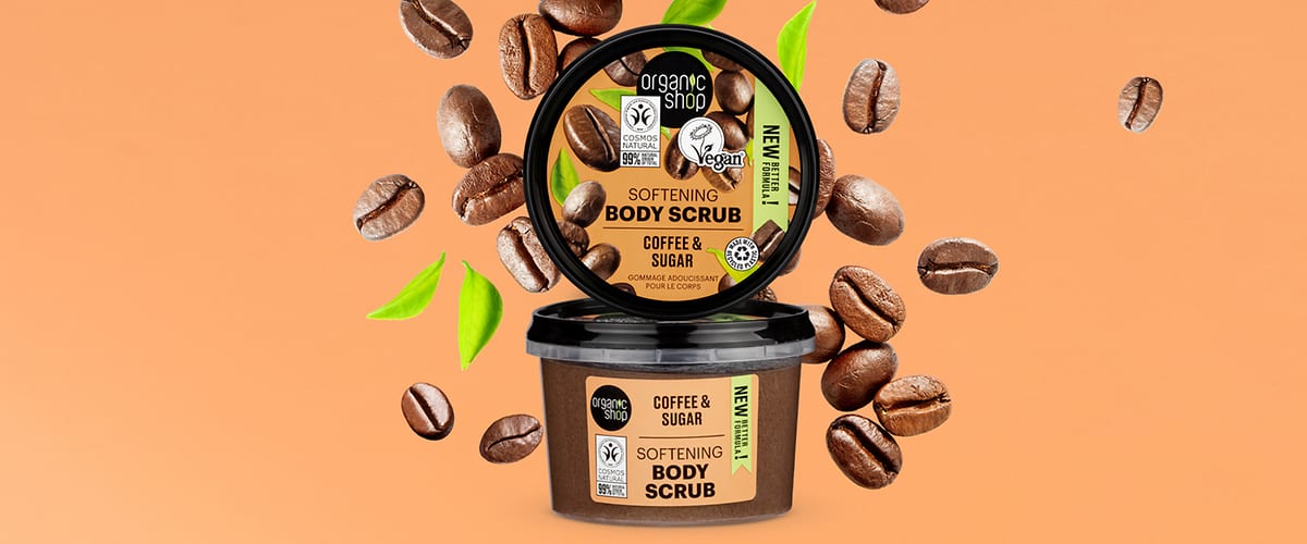 Recipient de scrub de corp cu cafea și zahăr, înconjurat de boabe de cafea și frunze verzi.