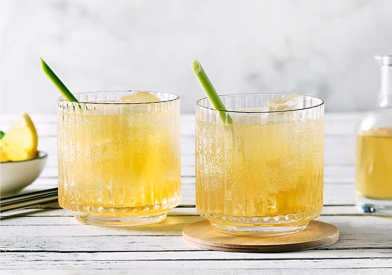 Două pahare de cocktail cu gheață și garnitură de lemongrass pe masă albă.