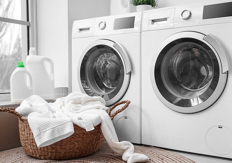 Două mașini de spălat albe lângă un coș cu rufe și detergent.