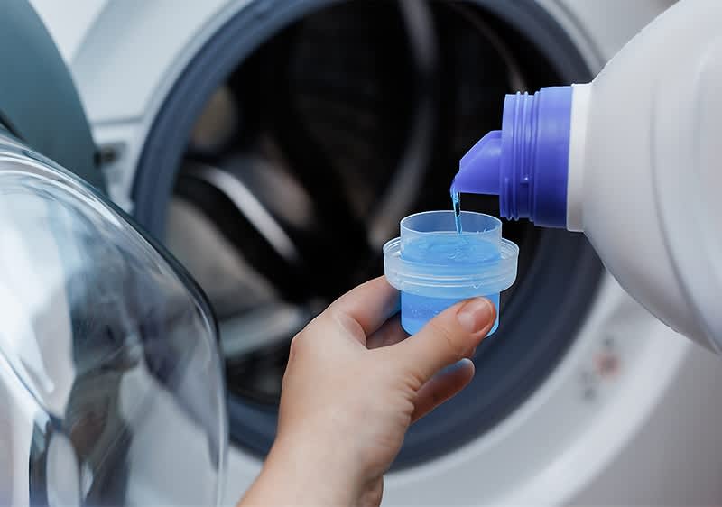 Persoană turnând detergent lichid într-un capac în fața unei mașini de spălat.