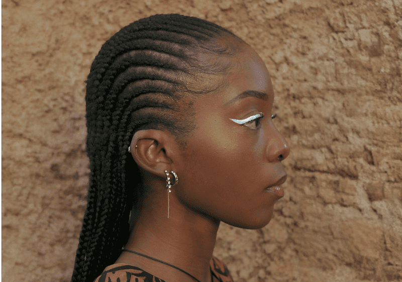 Persoană cu cornrows împletite lateral, cercel lung și fundal de perete de piatră
