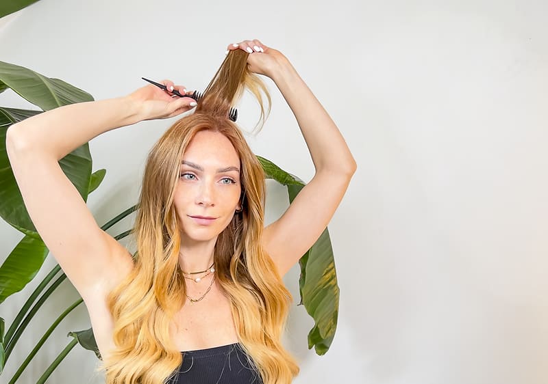 Femeie tânără coafându-și părul lung blond cu ondulare în fața unei plante decorative, brațe ridicate, fundal alb