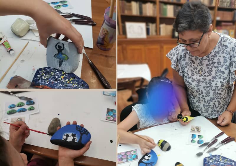 Pictură pe pietre în atelier cu persoane care pictează motive colorate pe pietre și modele expuse