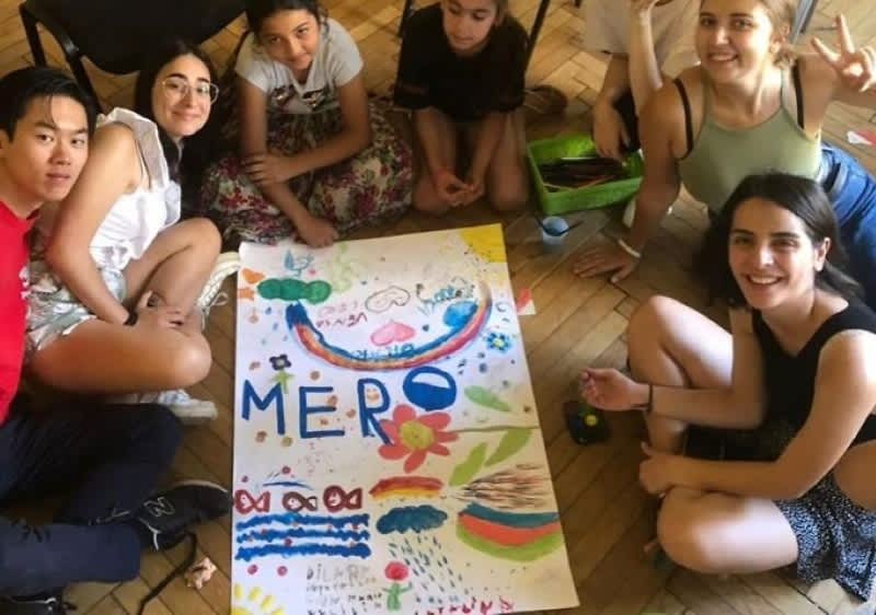 Copii așezați în cerc pe parchet în jurul unei picturi colorate pe hârtie intitulată MERO