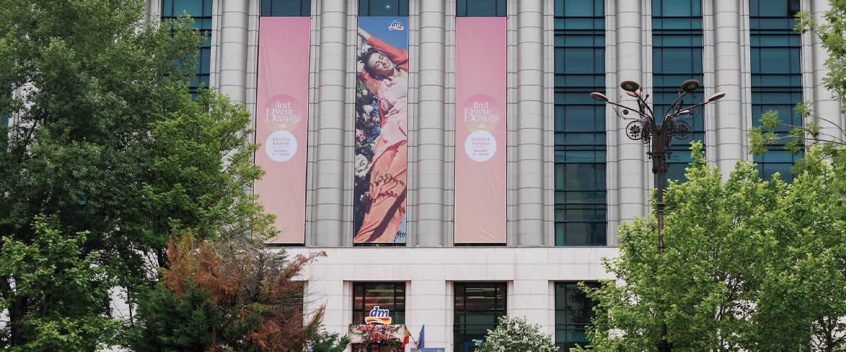 Fațadă de clădire cu trei bannere verticale roz și un banner central cu o femeie în rochie, ferestre și copaci în prim-plan