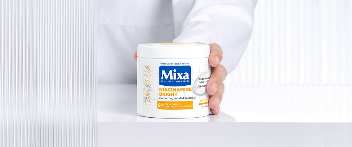 Recipientă albă de cremă Mixa Niacinamide Bright ținută de o mână pe fundal alb