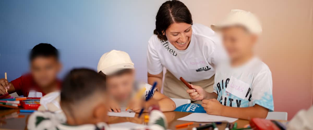 Activitate de desen cu copii la masă supravegheați de o femeie zâmbitoare