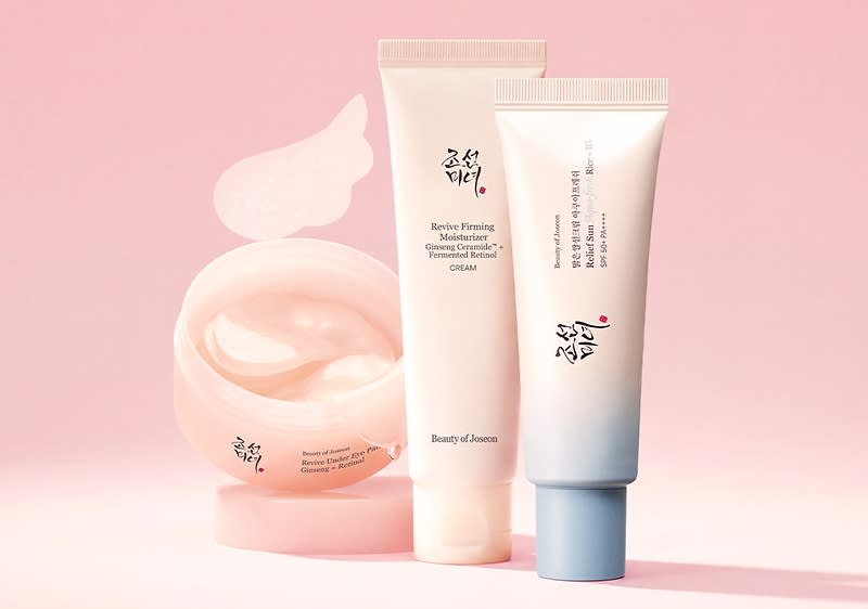 Un borcan roz deschis cu cremă deschisă la culoare lângă două tuburi verticale de produse cosmetice albe cu inscripții asiatice negre, unul cu capac roz pal și unul cu capac albastru deschis pe fundal roz pal