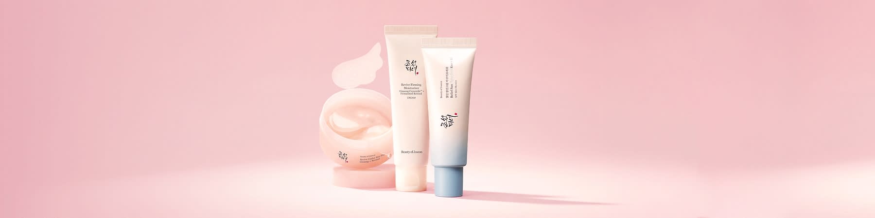 Trei produse cosmetice pe fundal roz: un borcan deschis cu cremă roz pal și două tuburi verticale, unul alb cu text „Revive Firming Moisturizer” și inscripții coreene, celălalt alb cu baze bleu și logo-ul mărcii Beauty of Joseon