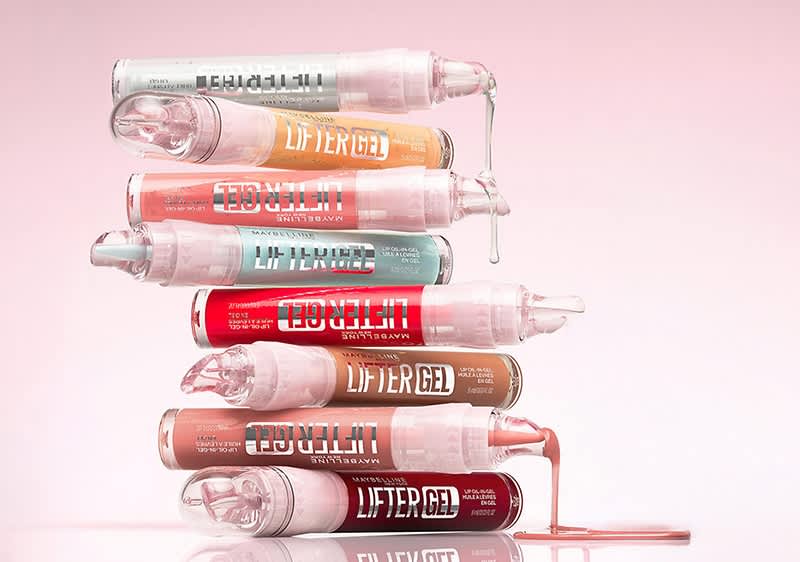 Optică de tuburi transparente de gloss stivuite orizontal în clăci; fiecare tub etichetat „LIFTER GEL” cu culori diferite de conținut: roz pal, piersică, corai, verde mentă, roșu, maro, roz intens și vișiniu, câteva picături gel curgând din tuburi pe fundal roz pal