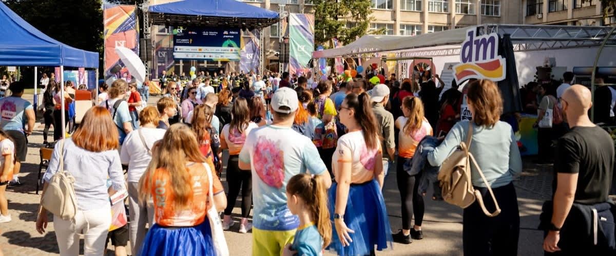 Mulțime de persoane la un eveniment în aer liber, majoritatea văzute de la spate purtând tricouri colorate și fuste albastre, scenă cu ecran și corturi de standuri în fundal, cort cu logo-ul „dm” vizibil pe partea dreaptă