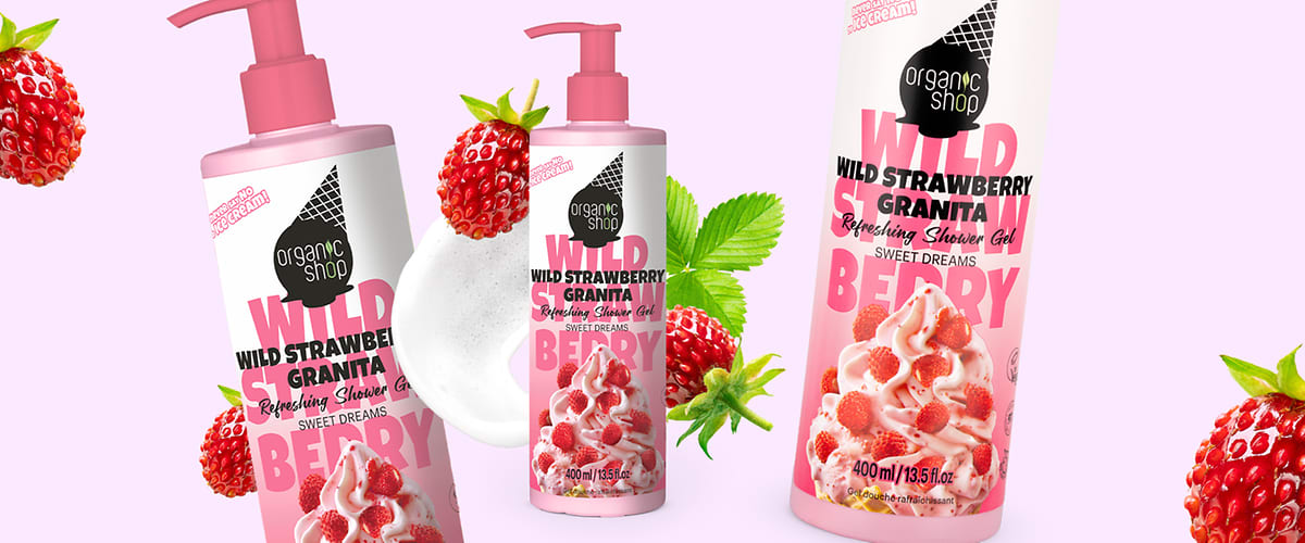 Trei flacoane roz cu pompă de gel de duș „Organic Shop Wild Strawberry Granita” cu etichetă albă și text roz și negru, imagini cu granita de căpșuni și fructe întregi, spumă albă și câteva frunze verzi pe fundal roz deschis