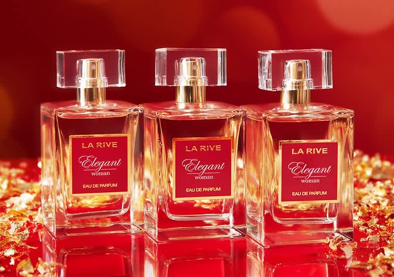 Trei sticle de parfum identice dispuse aliniat, sticlă pătrată transparentă cu lichid roz-auriu și dop pătrat transparent, etichetă roșie cu textul „LA RIVE Elegant woman Eau de Parfum” pe fundal roșu lucios cu sclipiri aurii
