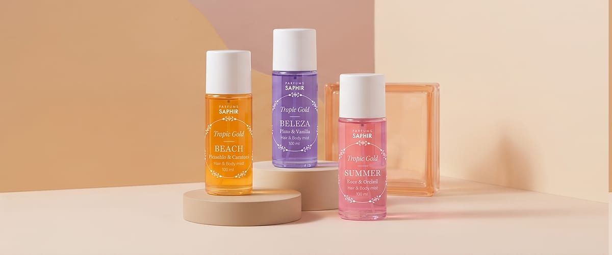 Sticlă roz transparentă de 100 ml cu etichetă albă pe care se citește „Parfums SAPHIR Tropic Gold SUMMER Rose & Orchid Hair & Body mist 100 ml”, alături de un recipient pătrat de sticlă portocaliu deschis, pe fundal crem