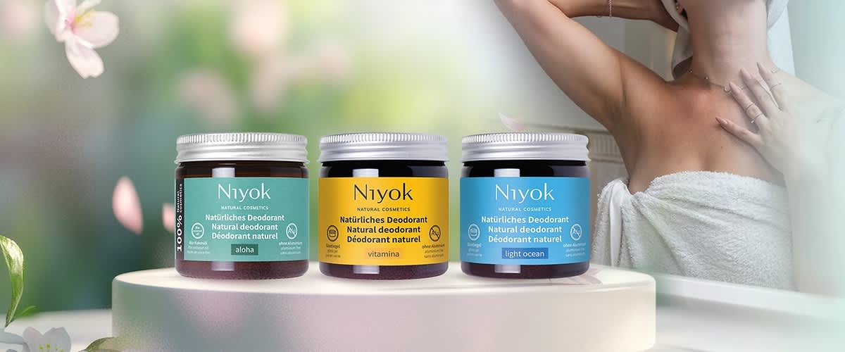 Trei borcane de deodorant solid Niyok aliniate pe o suprafață, de la stânga la dreapta etichete verzi „aloha”, galbenă „vitamina” și albastră „light ocean”, în fundal o femeie în prosop ridicând braţul pentru a-și arăta subraţul