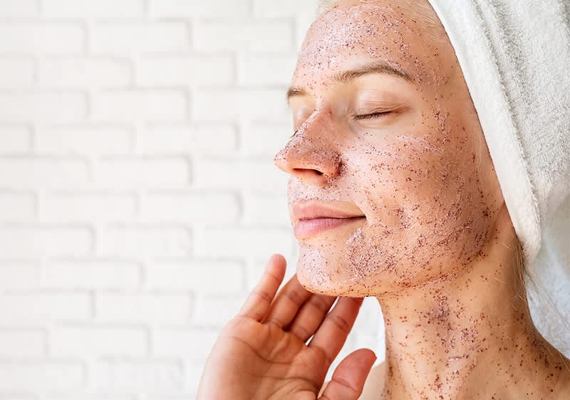 Osoba s uterákom na hlave nanáša peeling na tvár a krk, rukou priťahuje pleť bez výrazu tváre