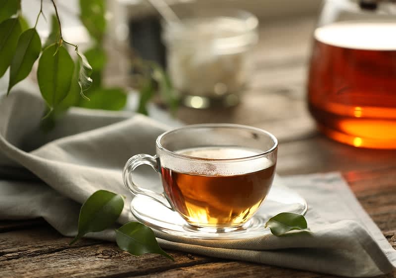 Šálka čierneho čaju na podšálke na látke s listami a kanvicou v pozadí