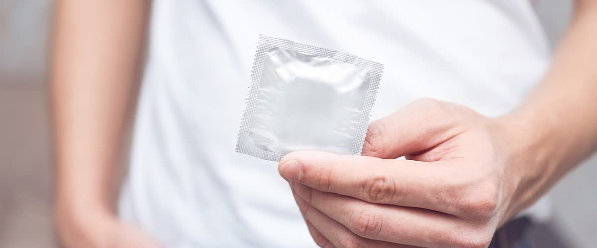 Muž s bielym tričkom drží v ruke zabalený kondóm