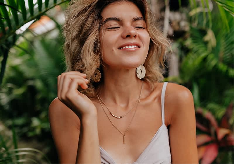 Žena v letných šatách s náhrdelníkom a náušnicami, obklopená zeleňou