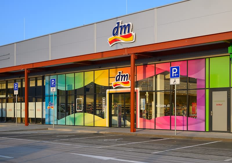 Exteriér predajne dm s farebnými sklenenými panelmi a parkovacími miestami pred budovou
