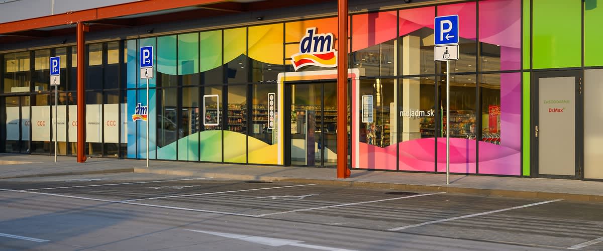 Exteriér predajne dm s farebnými sklenenými panelmi a parkovacími miestami pred budovou