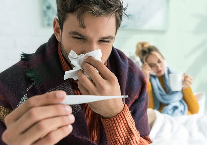Muž drží teplomer, v pozadí žena s hrnčekom v ruke
