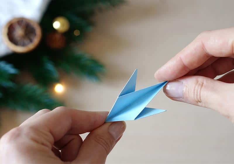 Zwei Hände falten ein blaues Origami-Papier zu einem Vogel, im Hintergrund sind Tannenzweige und eine getrocknete Orangenscheibe sichtbar