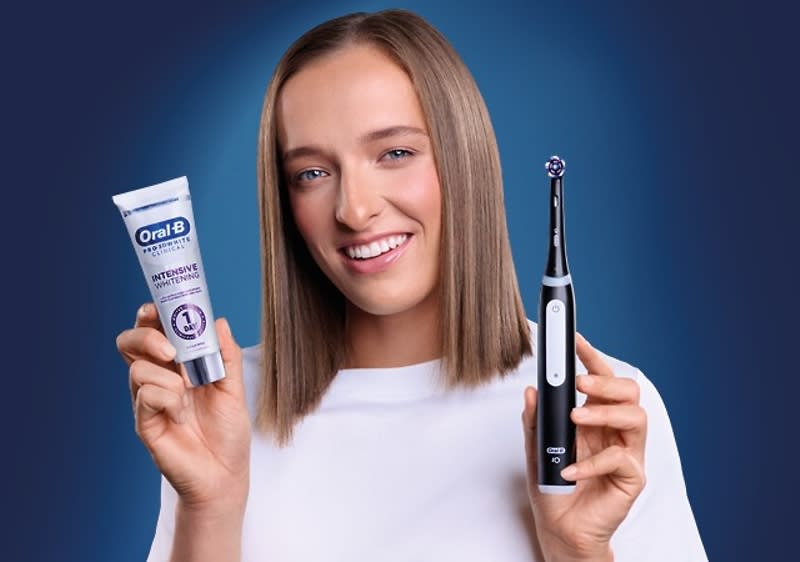 Osoba drží zubnú pastu Oral-B a elektrickú zubnú kefku Oral-B na modrom pozadí.