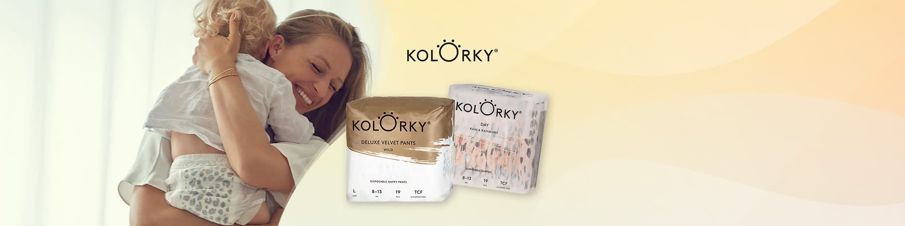 Matka objíma malé dieťa v plienkach, v popredí dve balenia jednorazových nohavičkových plienok značky Kolörky, jedno zlatobiele s nápisom „KOLÖRKY Deluxe Velvet Pants Wild“ a jedno bielo-ružové s nápisom „KOLÖRKY Day Rain & Rainbows“