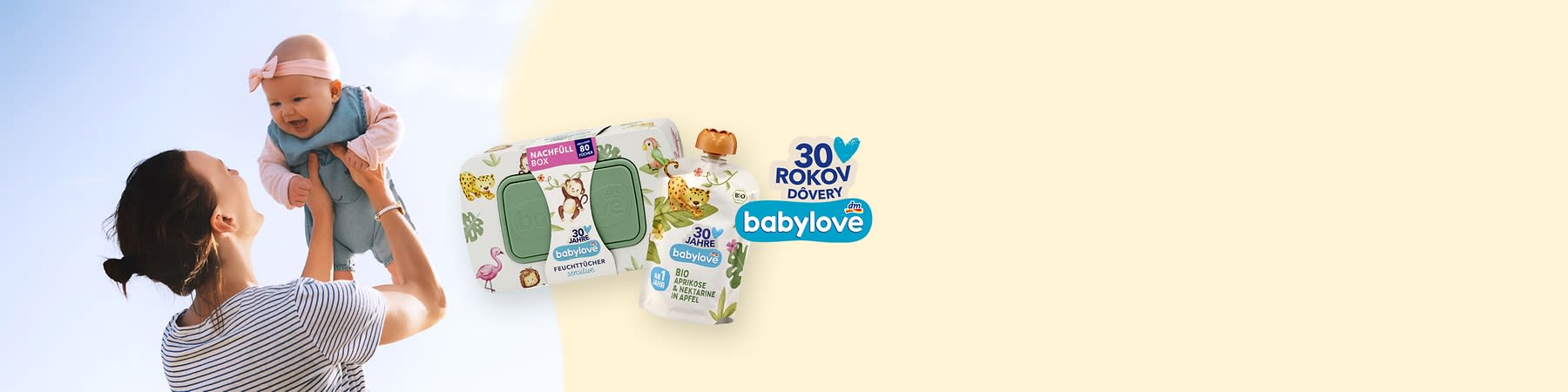 Matka zdvíha usmievajúce sa dojča v ružovej čelenke, vľavo viditeľná iba horná časť jej tela v pruhovanom tričku, vpravo balenie vlhčených obrúskov s zeleným viečkom a náhradné balenie kapsičky detského desertu s nápisom „30 rokov dôvery babylove“ a ilustráciami zvieratiek a ovocia