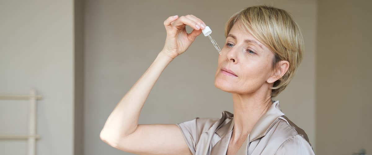 Žena stredného veku s krátkymi vlasmi nakláňa hlavu a kvapká si tekuté sérum