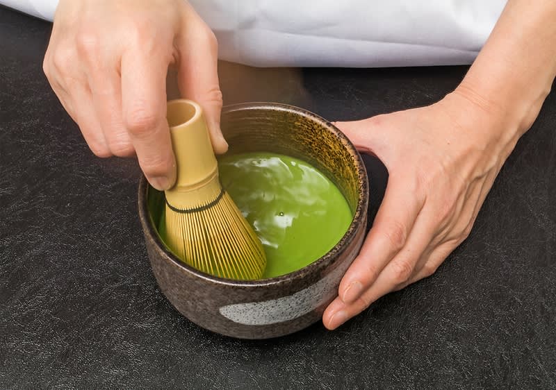 Ruky držia keramickú misku so svetlozeleným matcha čajom, jedna ruka pridŕža misku, druhou sa bambusovým metličkovým šľahačom mieša pena v nápoji