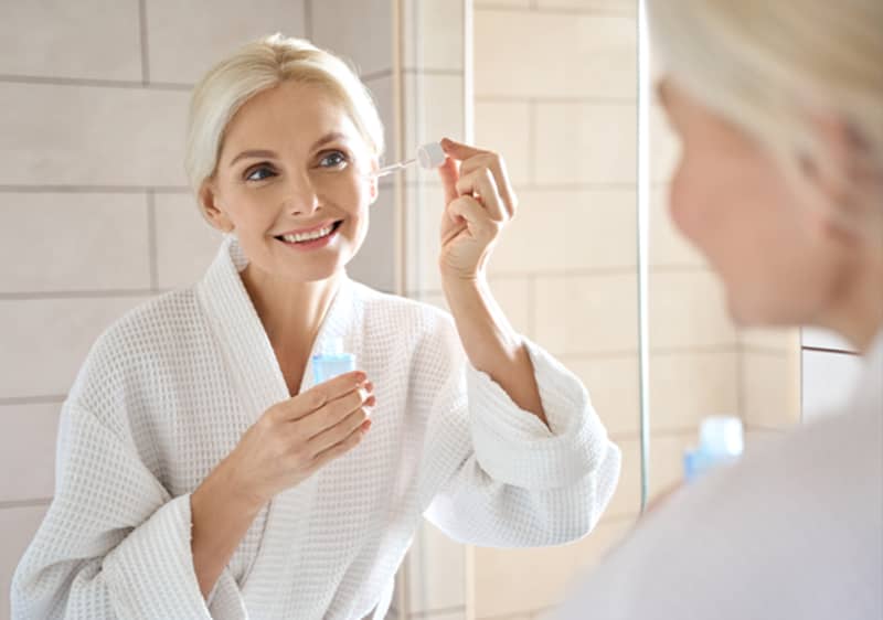 Žena v župane si nanáša sérum pred zrkadlom.