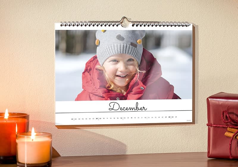 Kalendár na stene s obrázkom dieťaťa v zimnom oblečení, vedľa sviečky a darčekovej krabičky