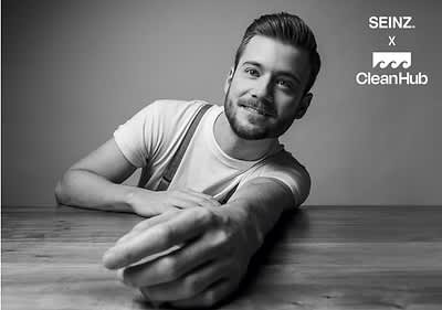 Moški v beli majici sedi za mizo z iztegnjeno roko, zamegljen obraz, logotipi SEINZ. in CleanHub