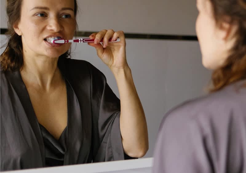 Oseba v kopalnem plašču si pred ogledalom ščetka zobe