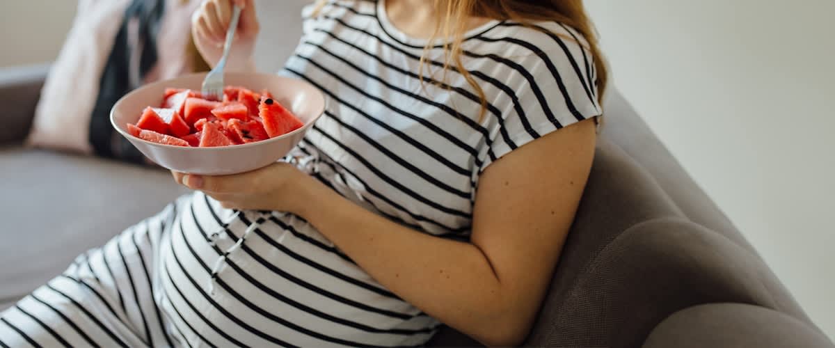 Nosečnica v črtasti majici je na kavču in je lubenico.