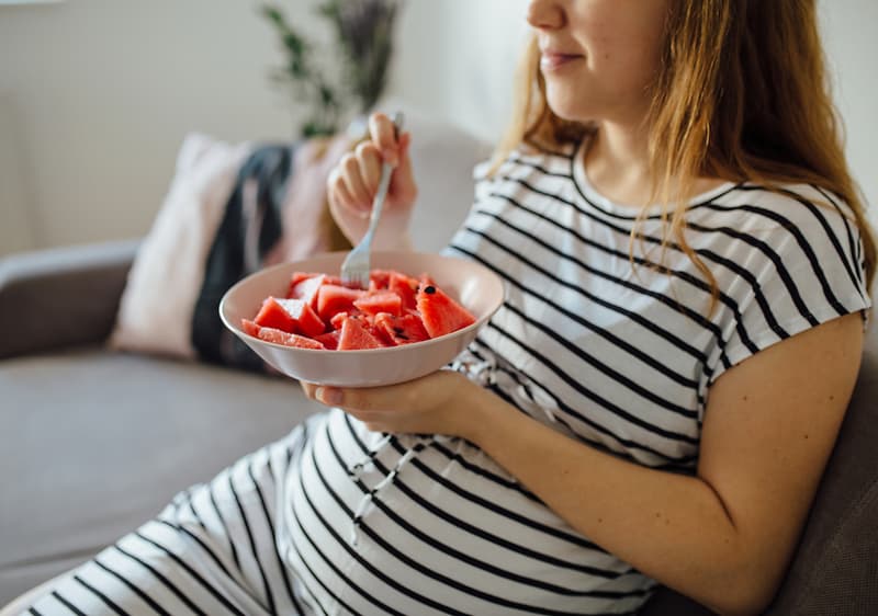 Nosečnica v črtasti obleki je na kavču in je lubenico iz sklede.
