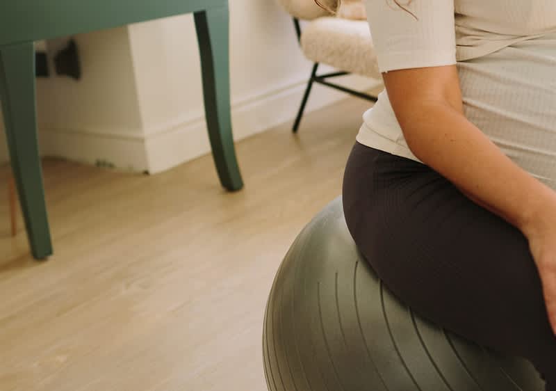 Nosečnica sedi na vadbeni žogi v dnevni sobi.