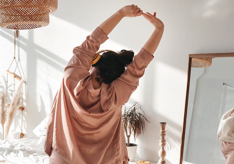 Ženska v pižami se razteza v spalnici z ogledalom in rastlino.