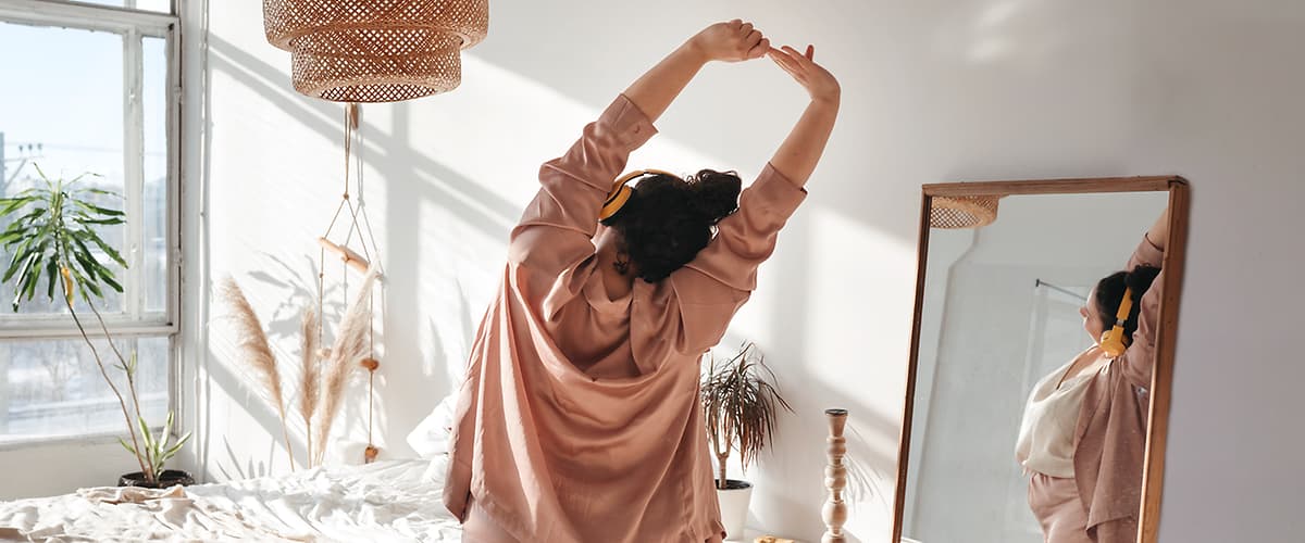 Ženska v pižami se razteza v spalnici z ogledalom in rastlino.