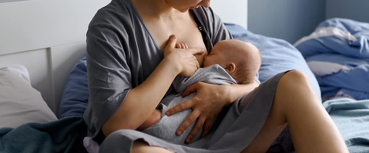 Dojenje dojenčka v naročju matere v sivi obleki na postelji